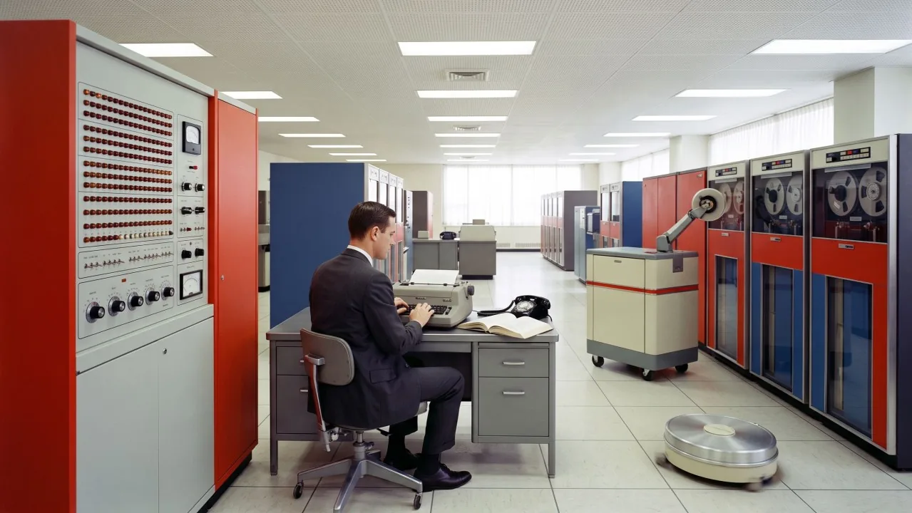 A machine room that never existed, 1965. The narrow machines do the narrow work; the person is still deciding. Image generated with AI.